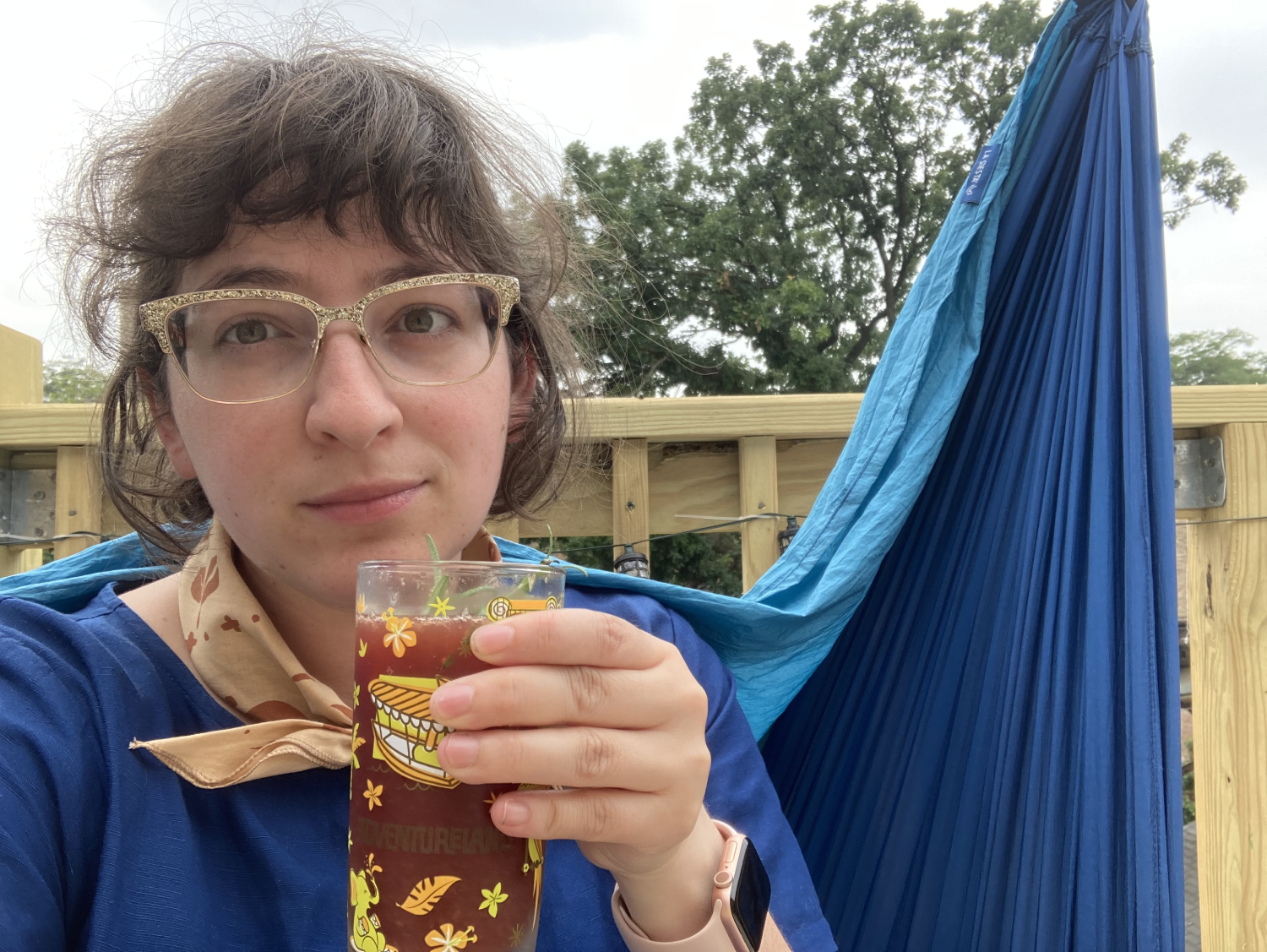 Camp Director Summer wearing a blue shirt and yellow bandana, sitting on her porch enjoying her pom cocktail
