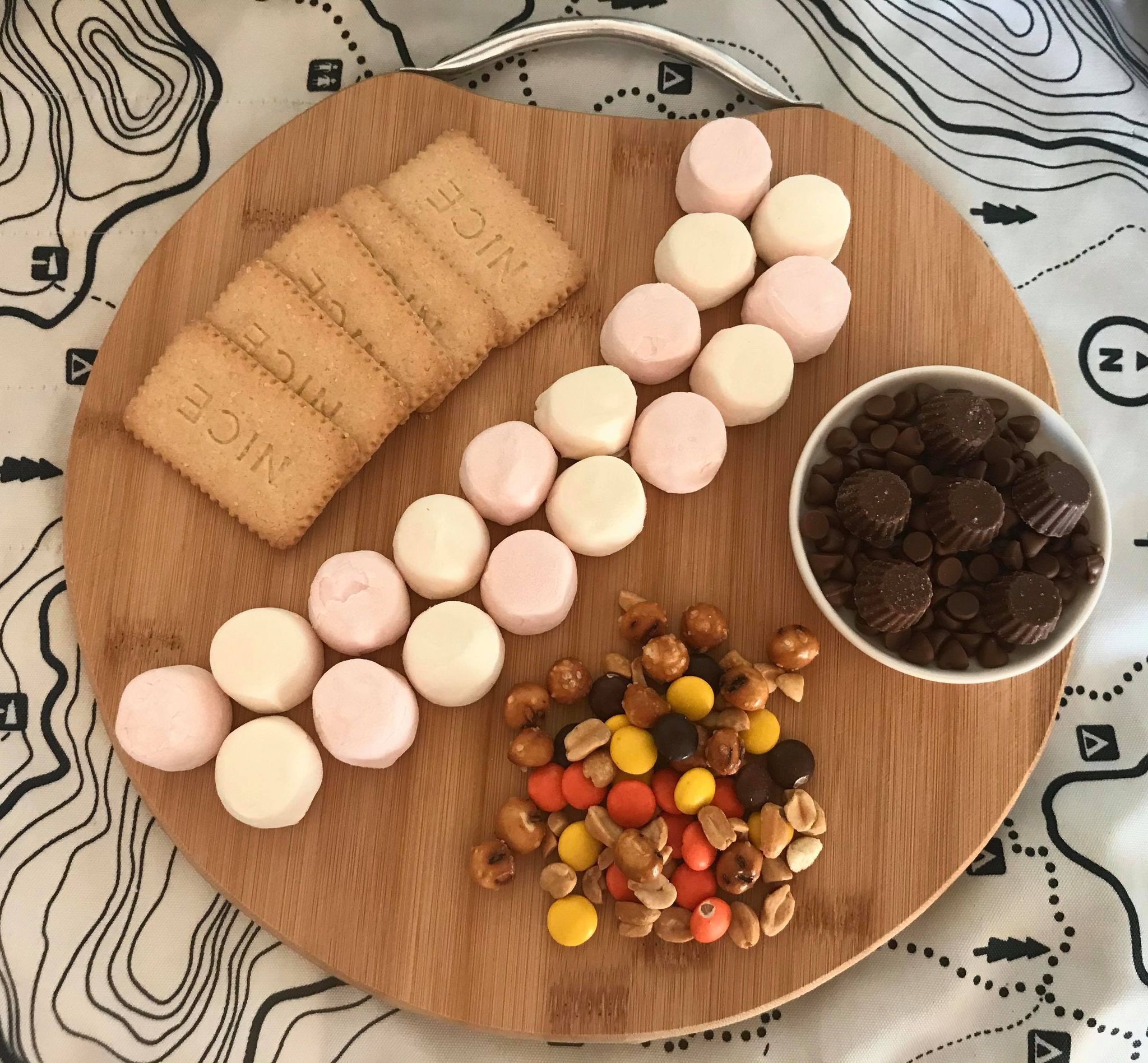 a round wooden board with biscuits, marshmallows, small candies, and chocolate dipping sauce arranged on it