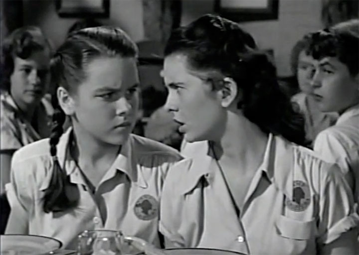 Still from the 1951 film Her First Romance, with two young girls wearing camp uniforms and intently conversing.