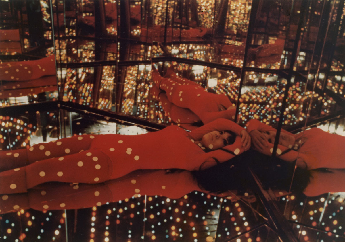 A photograph of the Japanese artist, Yayoi Kasuma, wearing a red body suit covered in dots, in a mirror room, one her exhibition installations.