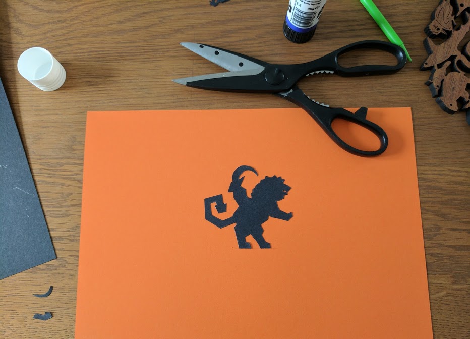 Chimera silhouette cutout in the center of a large piece of orange cardstock, with a pair of scissors beside it