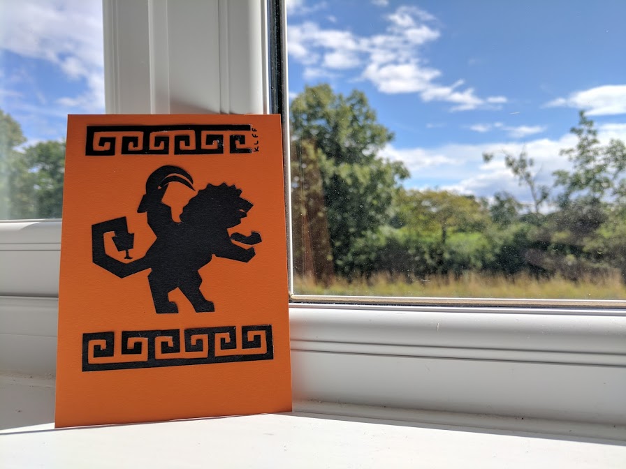 final cut out version of the postcard sitting on a windowsill; chimera creature in the center of the postcard with the geometric pattern above and below