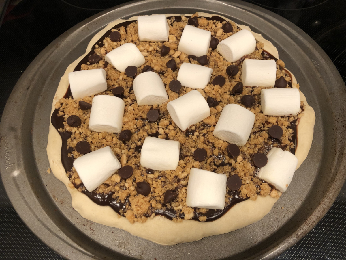 Unbaked s'mores pizza with a base of pudding, then crushed graham, then chocolate chips and marshmallows spread evenly across the top