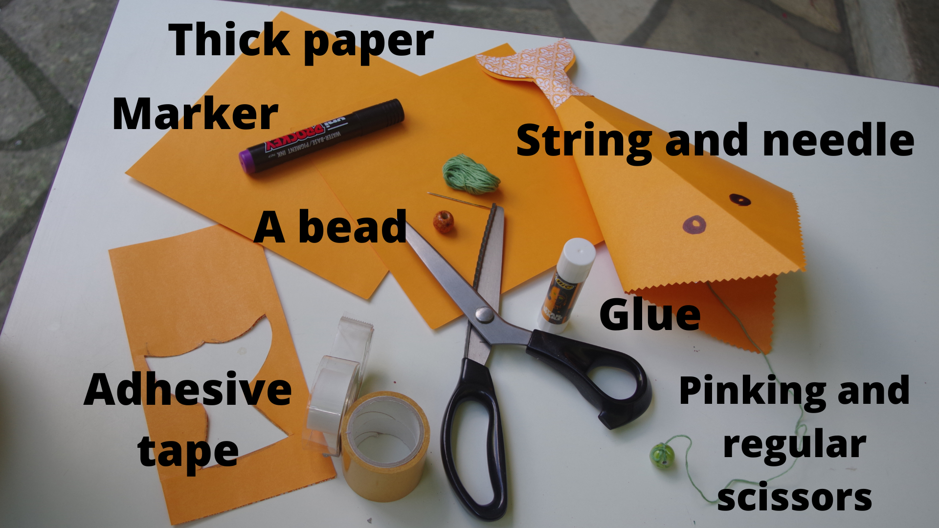 Supplies for the project are arrayed on a white surface, including orange paper, a pair of scissors, a bear, string with a large needle, adhesive tape, glue, and a marker