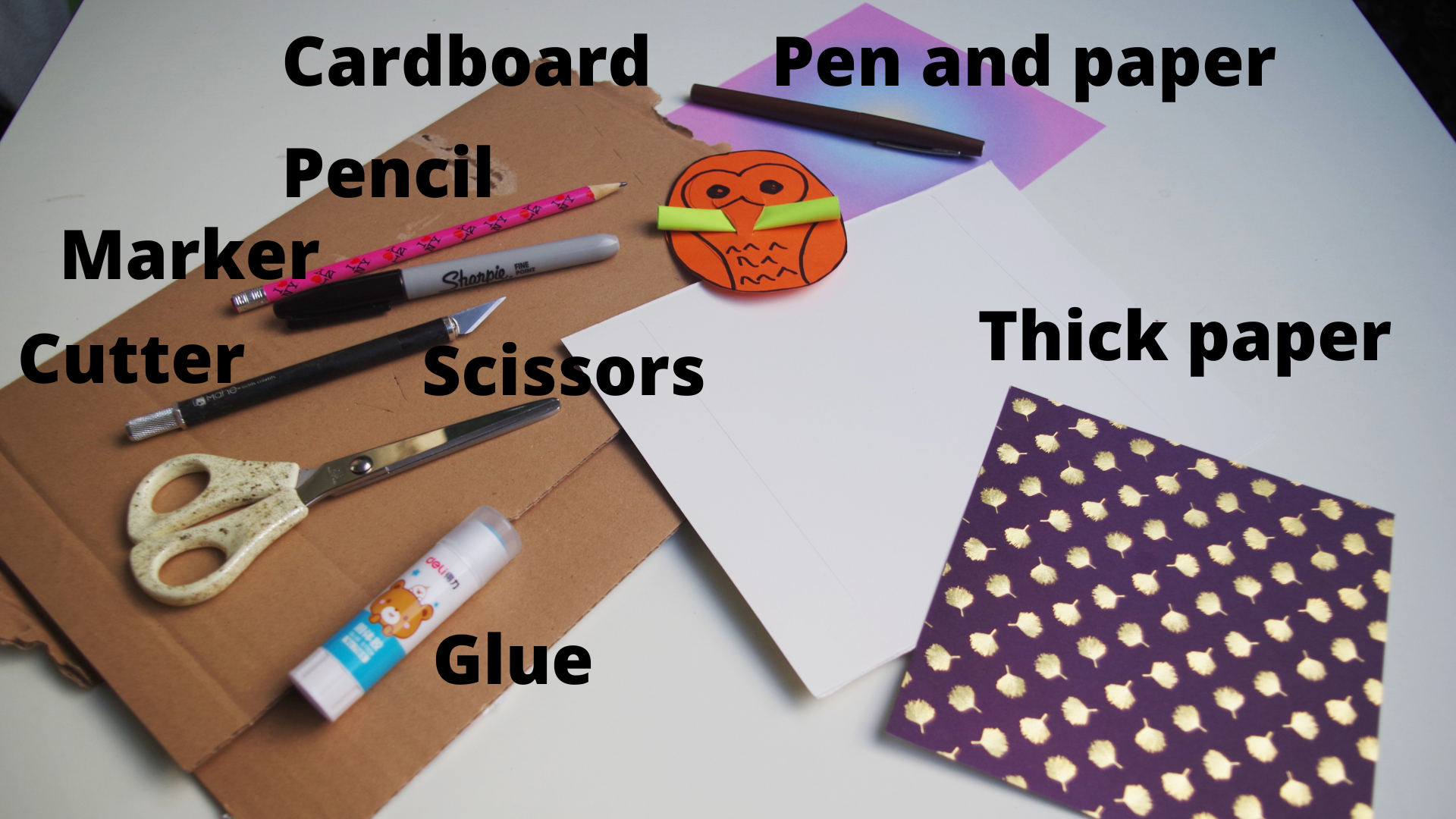 Supplies to make custom secret message owl cards arranged over a surface. Supplies include cardboard, pen and paper, thick crafting paper, glue, scissors, markers, and something to cut with