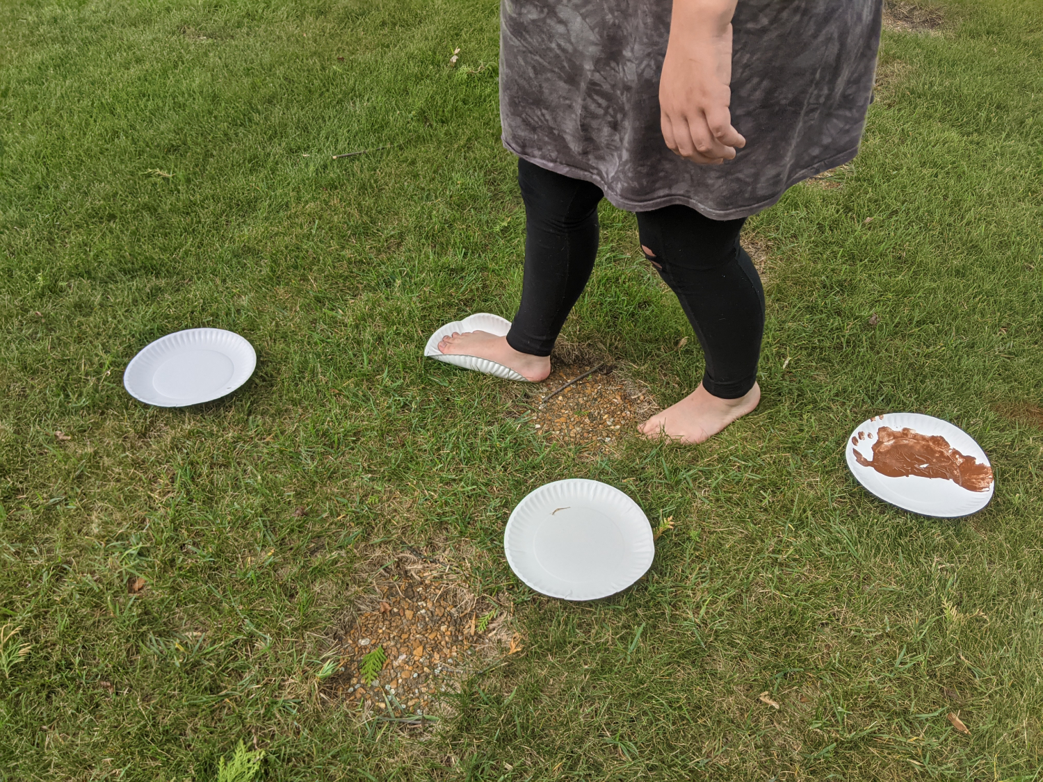 Paper plates are arranged in an alternating line (like footsteps) on a lawn. A person with brown paint on the bottom of their feet carefully steps from plate to plate leaving painted footprints behind. 