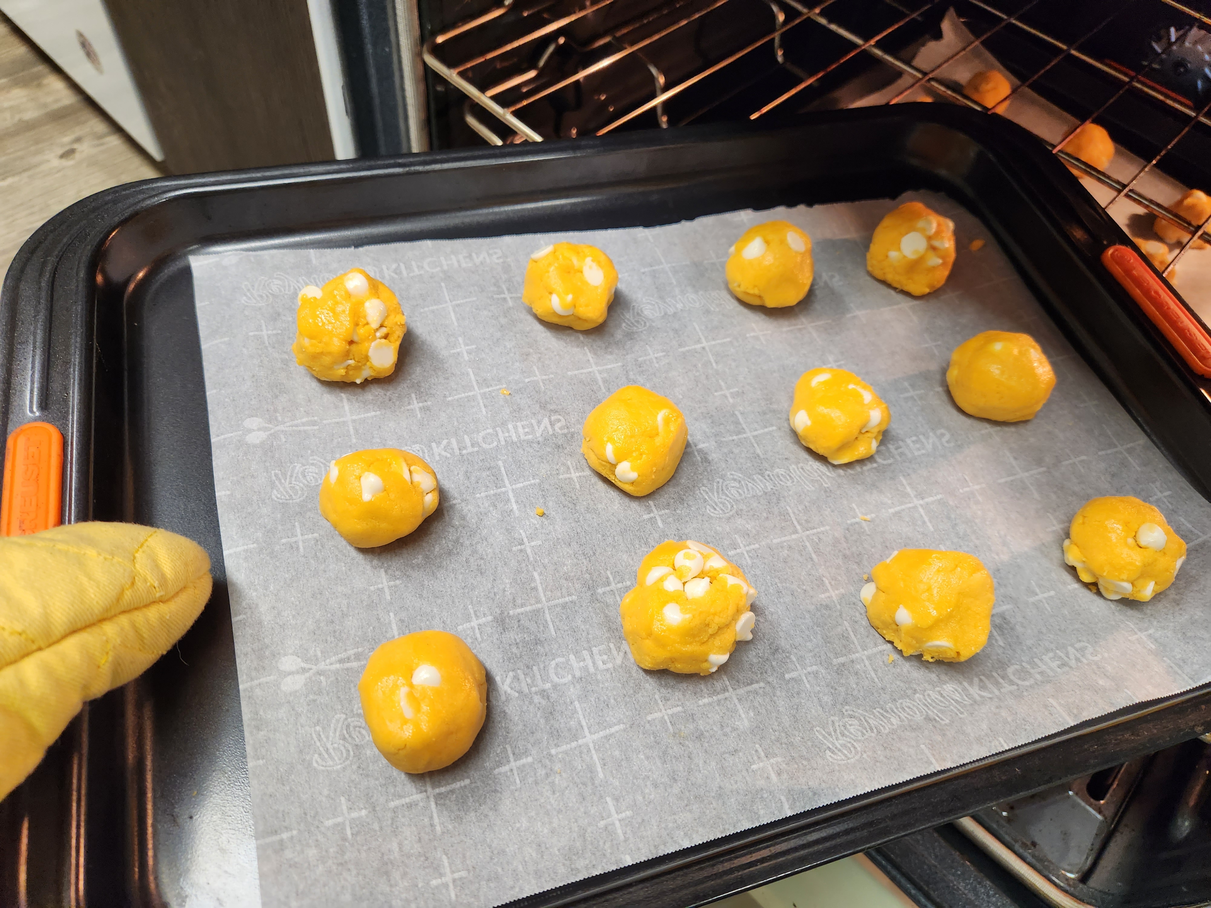 a dozen dough balls evenly spaced on a baking sheet being slid into the oven using a light yellow oven mitt