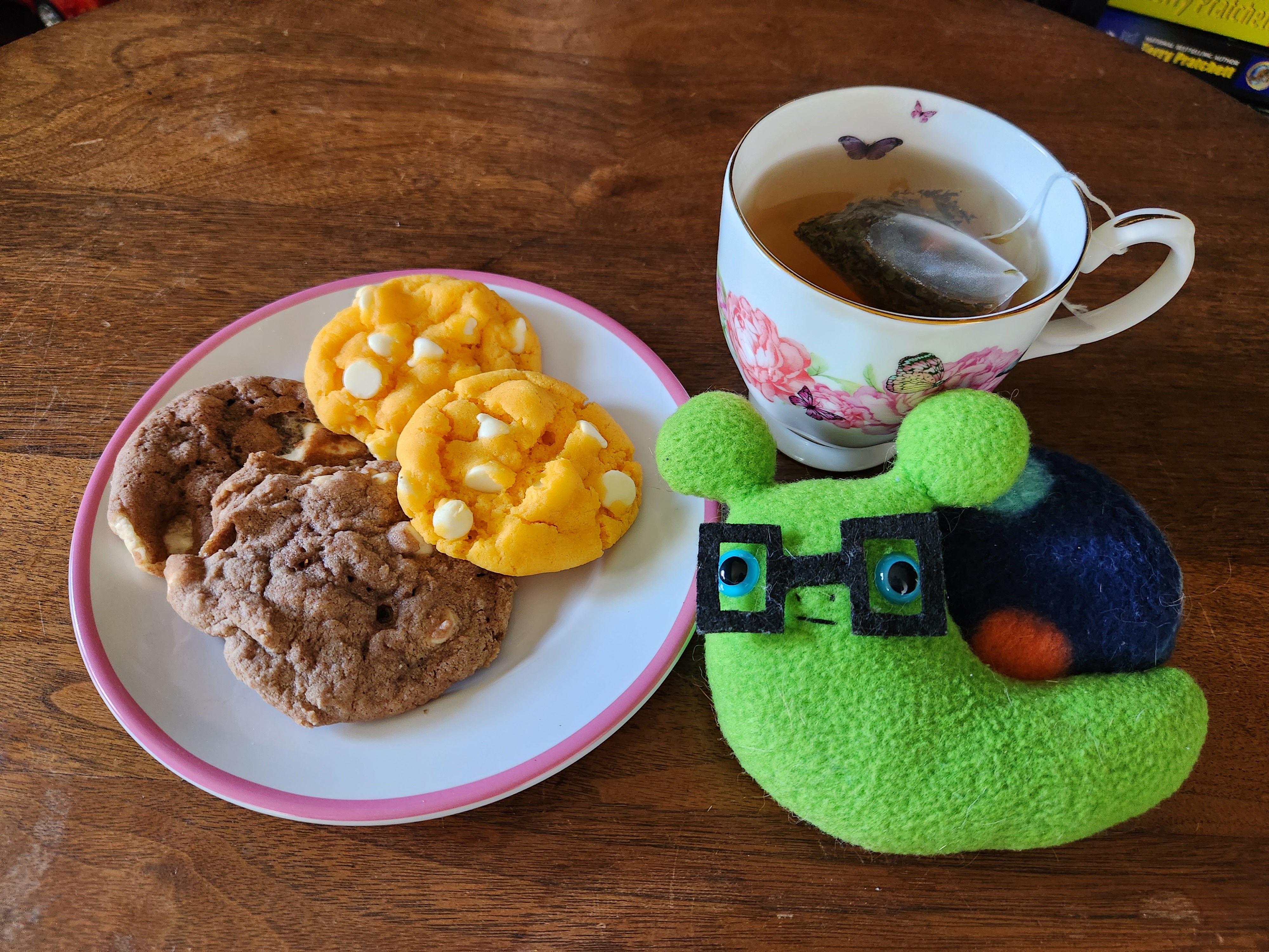 a plate of four cookies, two orange and two chocolate, sit on a wooden table with a cup of tea brewing and a little plush JimBob Snail