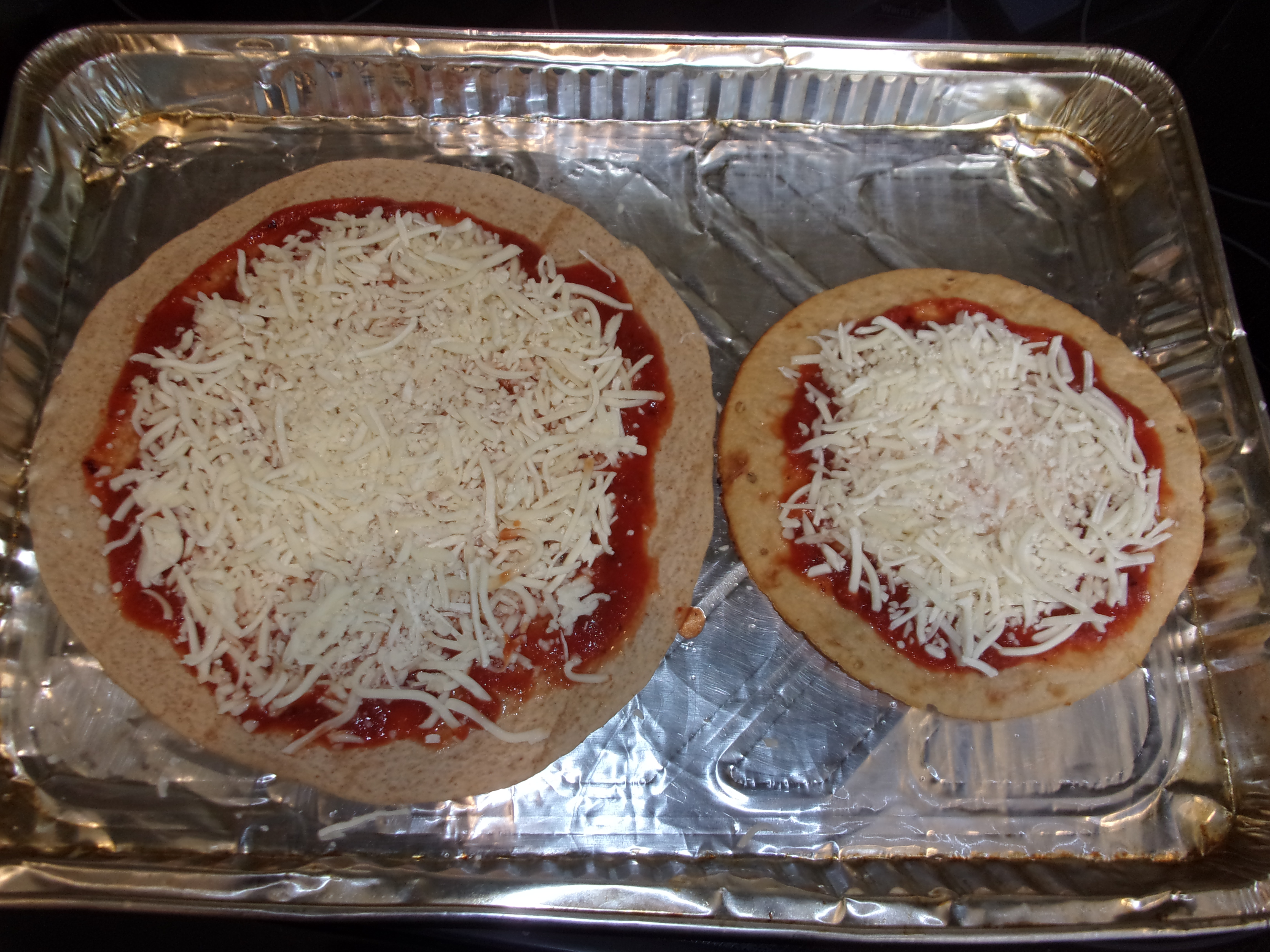 two different pizza bases on foil: one is a tortilla, one is a mini pizza crust. both have red sauce and shredded cheese spread across them