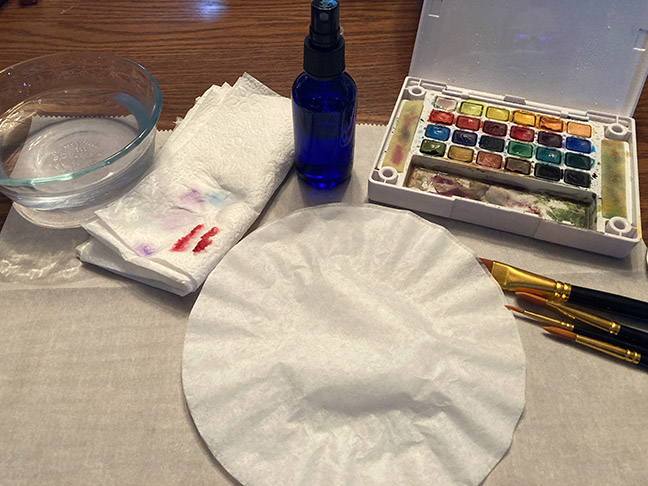 supplies needed for this project including a coffee filter, a spray bottle, watercolor paints, brushes, and a bowl of water