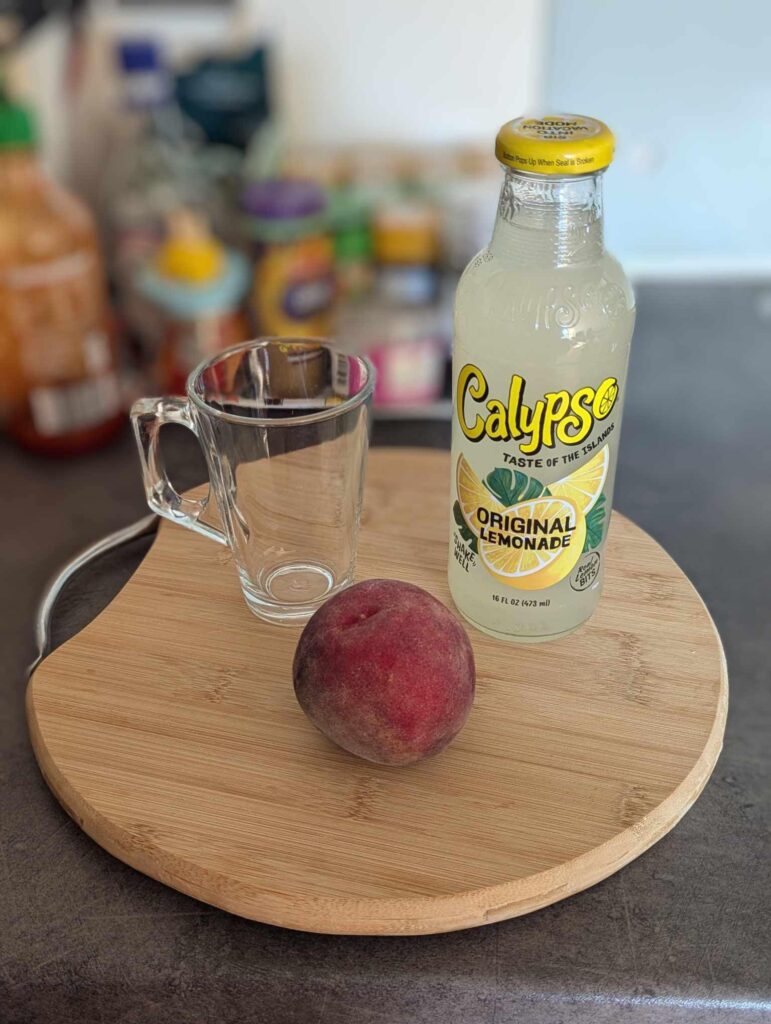 a glass mug, a bottle of lemonade, and a peach on a cutting board