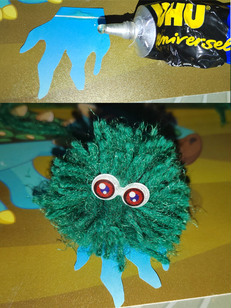 a green pompom with eyes and feet stands on a gold paper surface