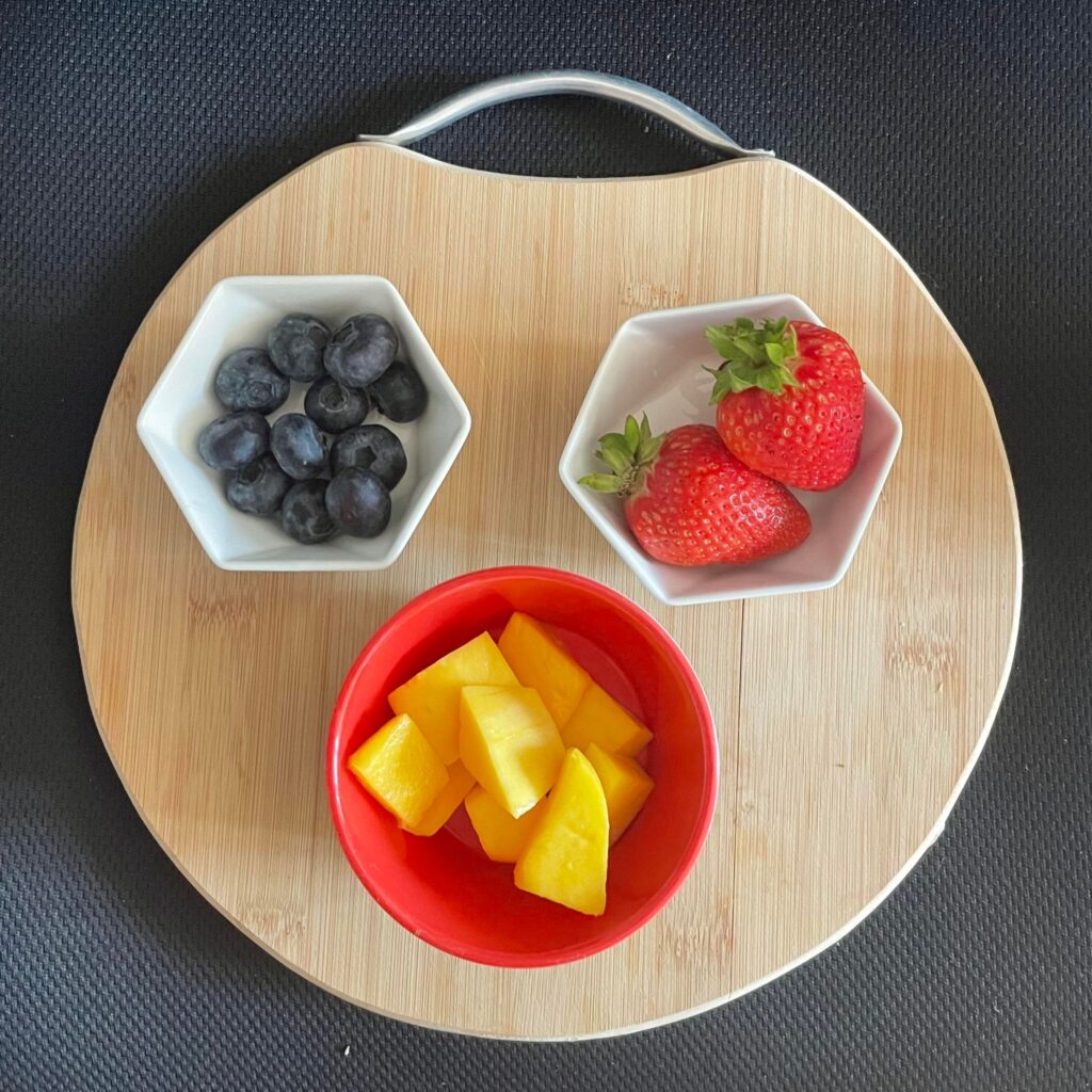 strawberries, blueberries, and mango cut and placed in small bowls on a cutting board