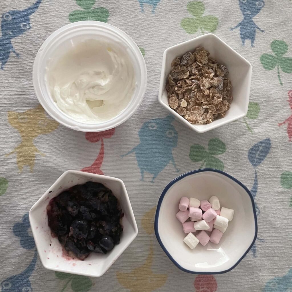 four small bowls containing yoghurt, marshmallows, chocolate chips and granola